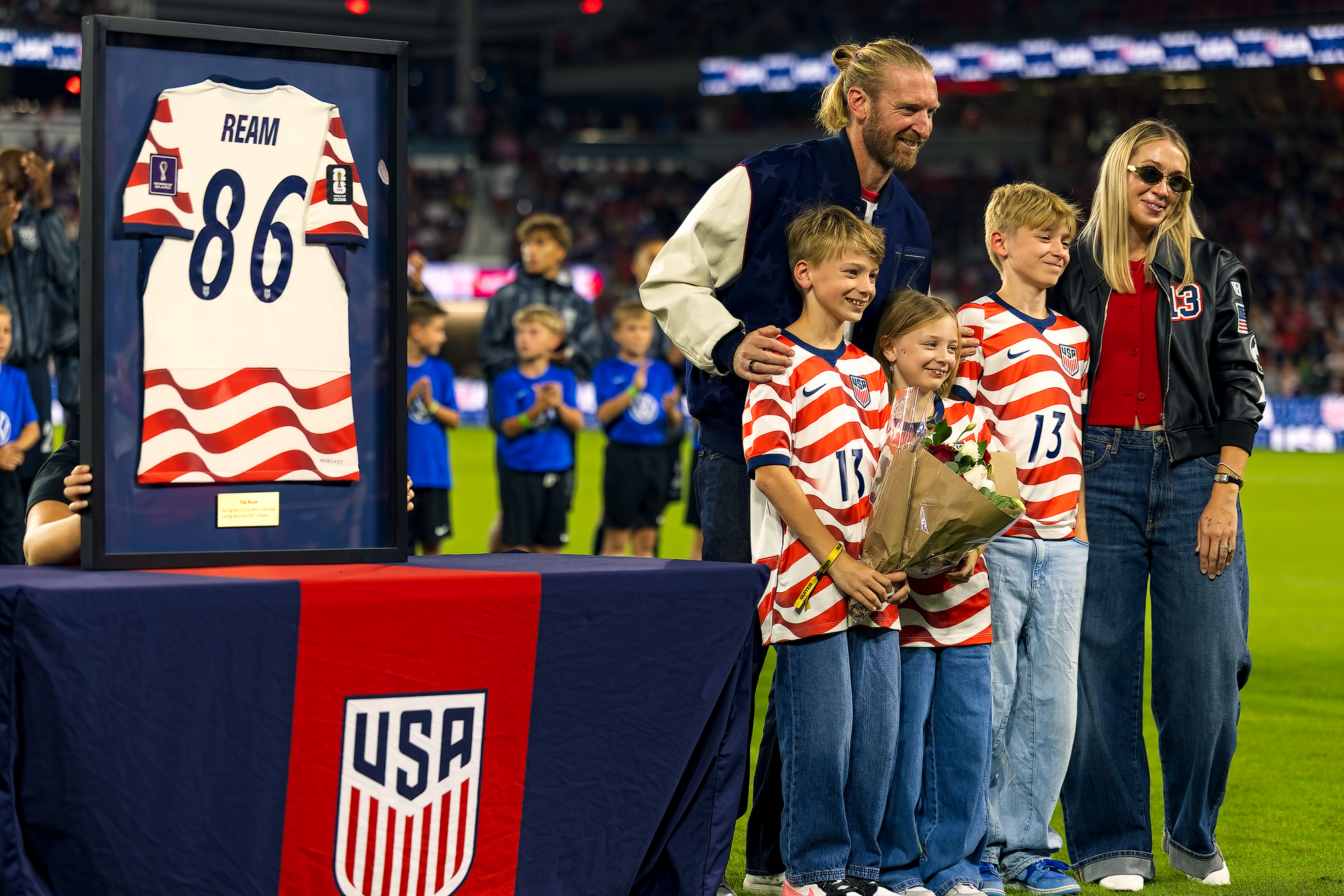 Tim Ream stands with his three children and his wife on the field beside a framed United States jersey bearing the number 86, displayed on a table with the U.S. Soccer crest, during a pregame retirement ceremony.