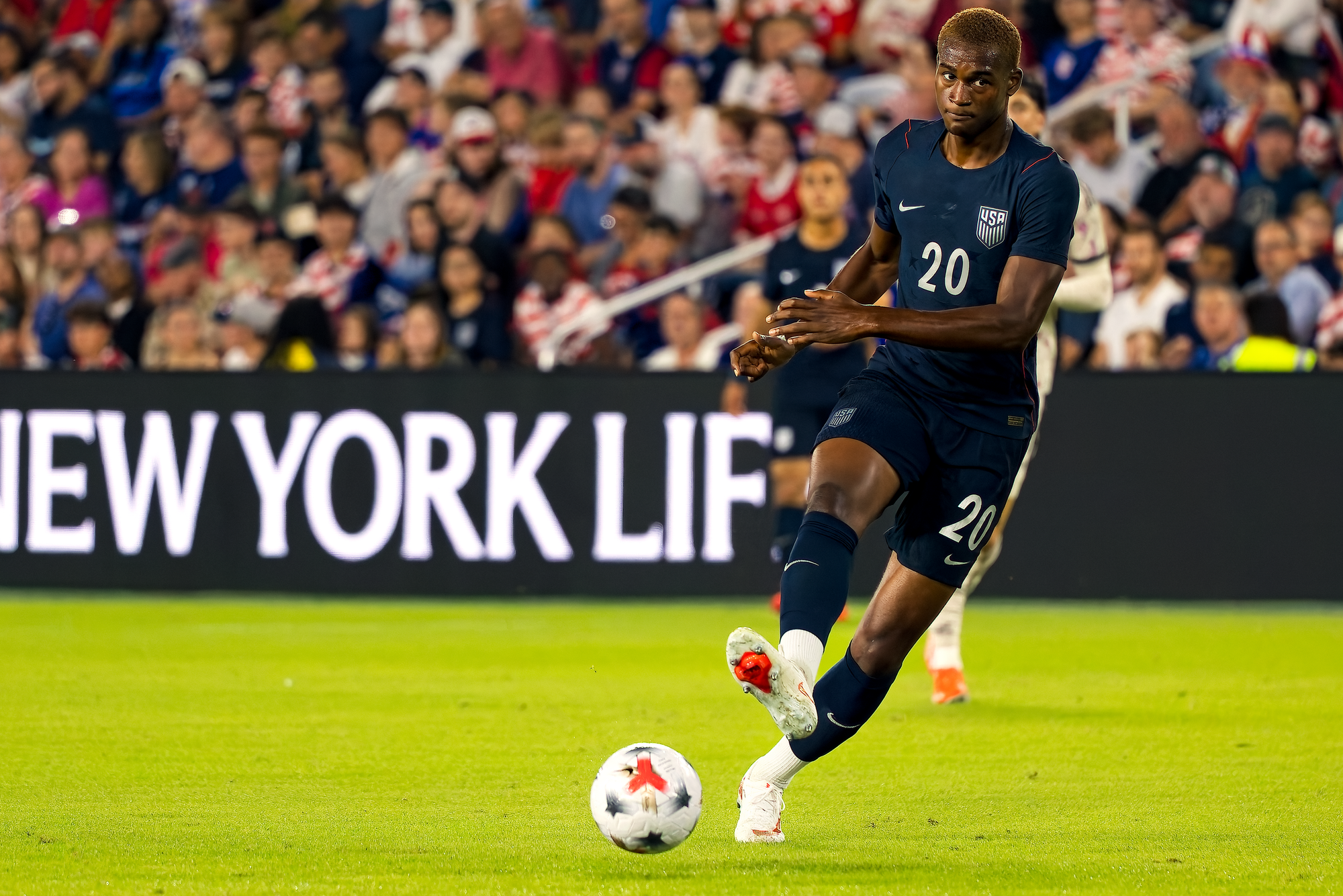Two United States players in navy kits converge on a Chile player in a white kit who is driving forward with the ball, with the referee and the goal behind them.