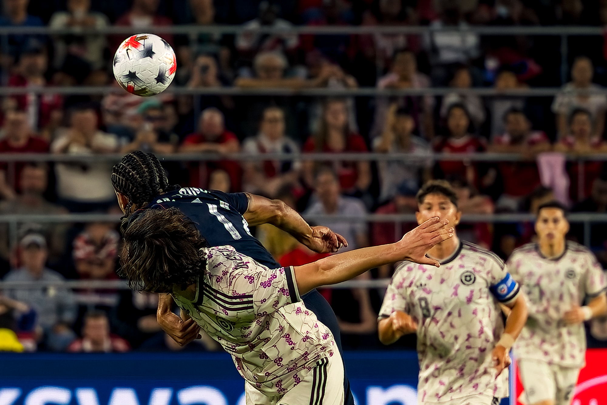 A United States defender and a Chile forward collide in midair competing for a header, with the ball suspended above them against a dark crowd.