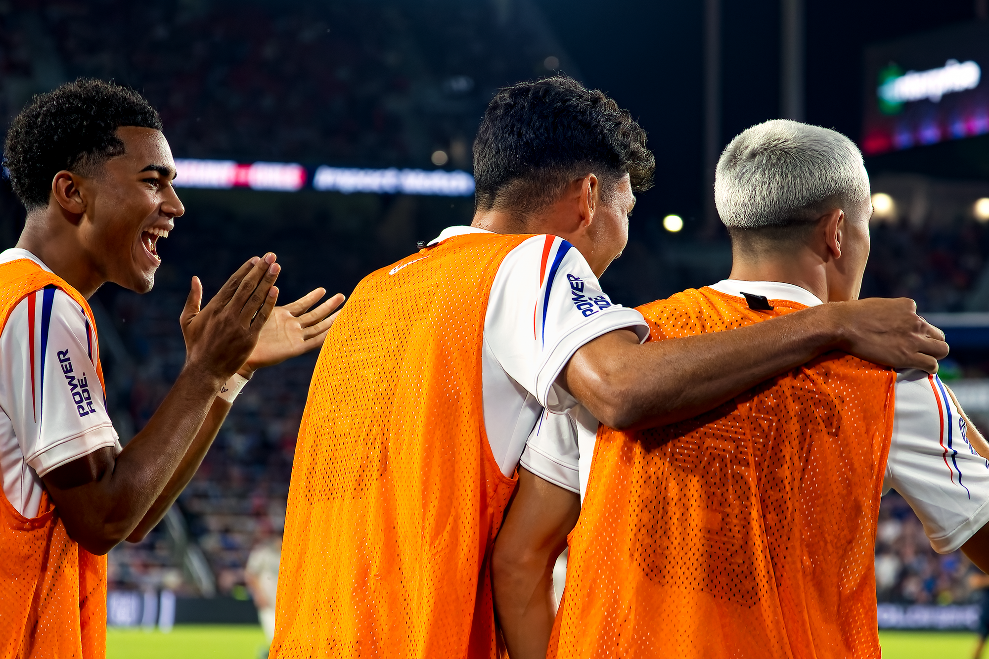 United States substitutes Zavier Gozo, Justin Ellis and Cavan Sullivan, in orange training bibs, laugh and embrace on the sideline while celebrating a goal.
