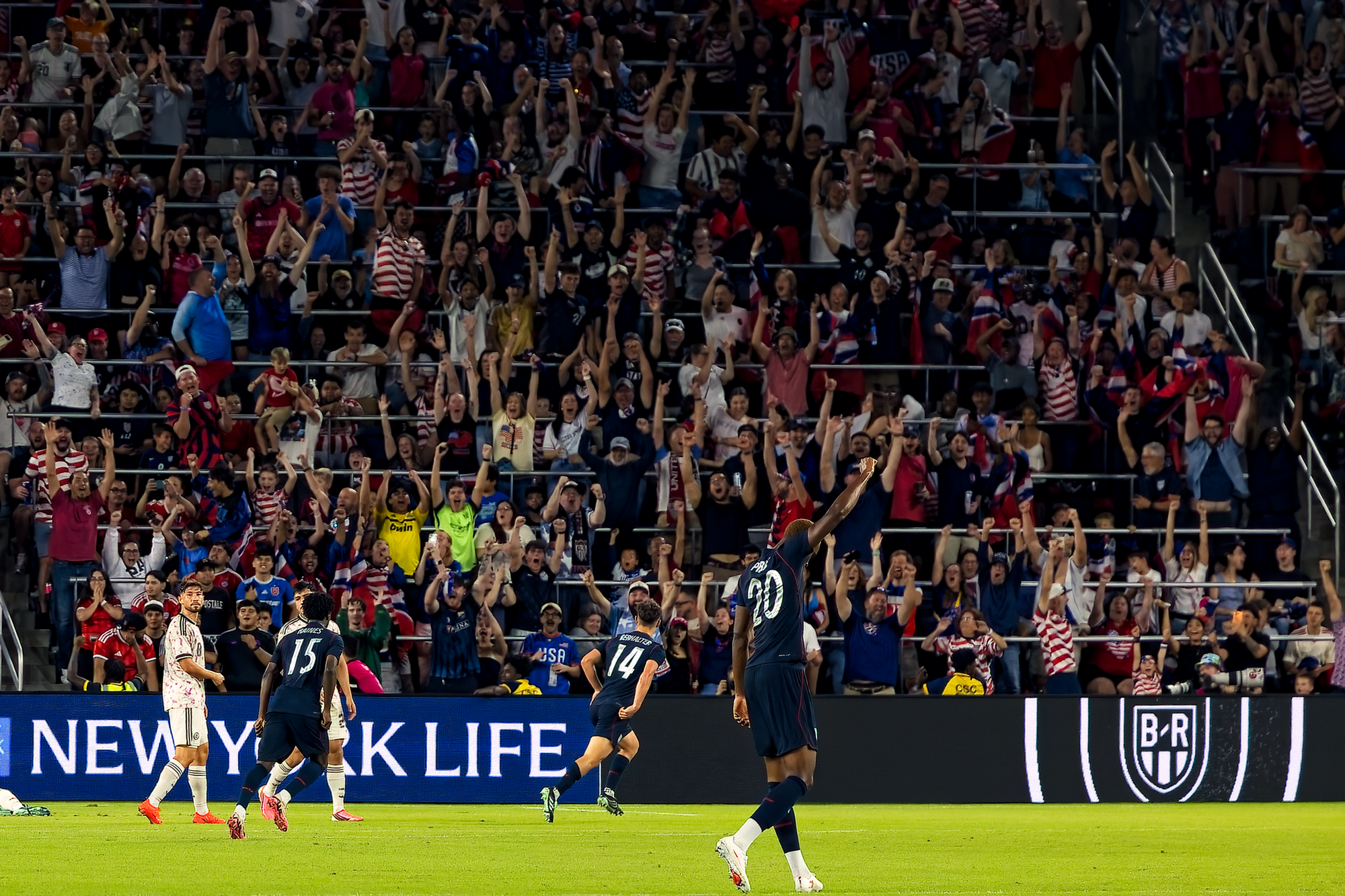 A United States player raises a fist in celebration as teammates run past and a packed stand of fans rises with arms in the air behind them.