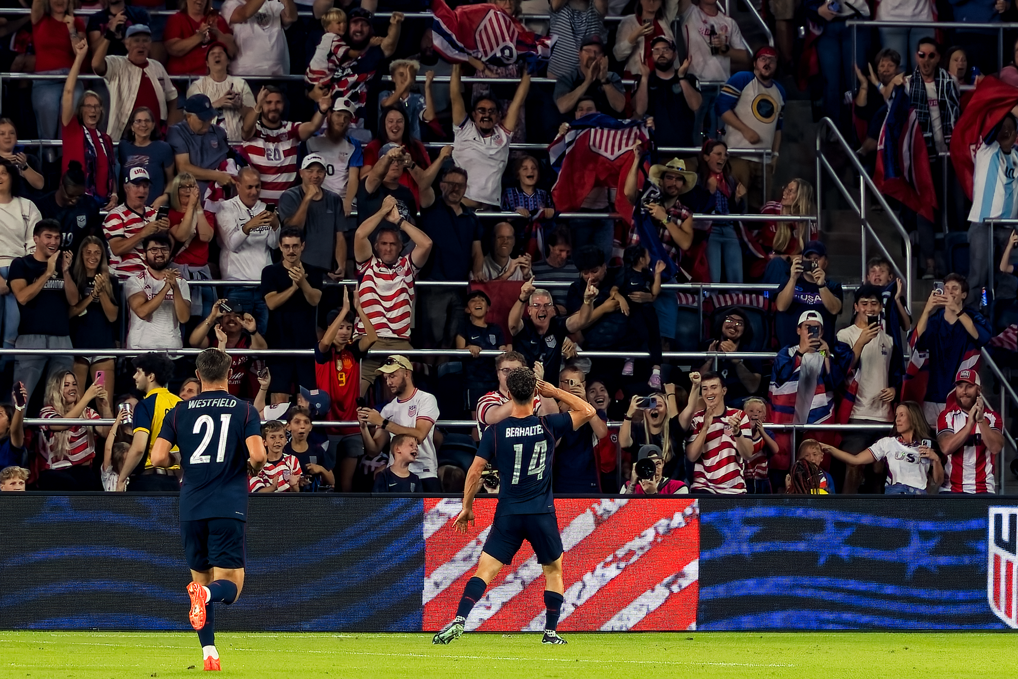 United States midfielder Sebastian Berhalter raises a hand to the crowd in celebration after scoring as a teammate runs toward him and fans cheer behind the advertising boards.