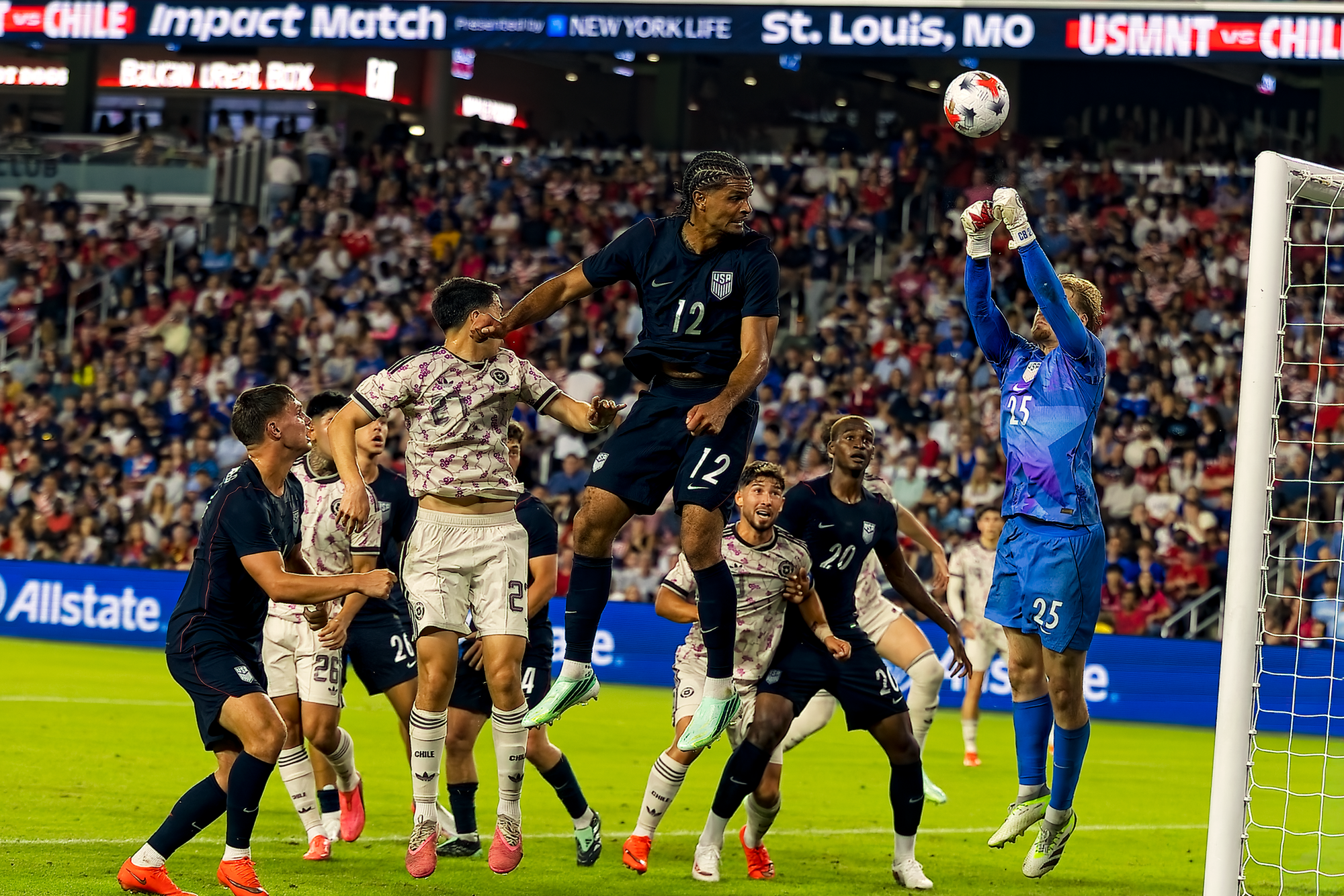 A United States goalkeeper in a blue kit stretches above a crowded six-yard box to parry the ball as a teammate leaps to challenge, with the St. Louis scoreboard banner behind them.