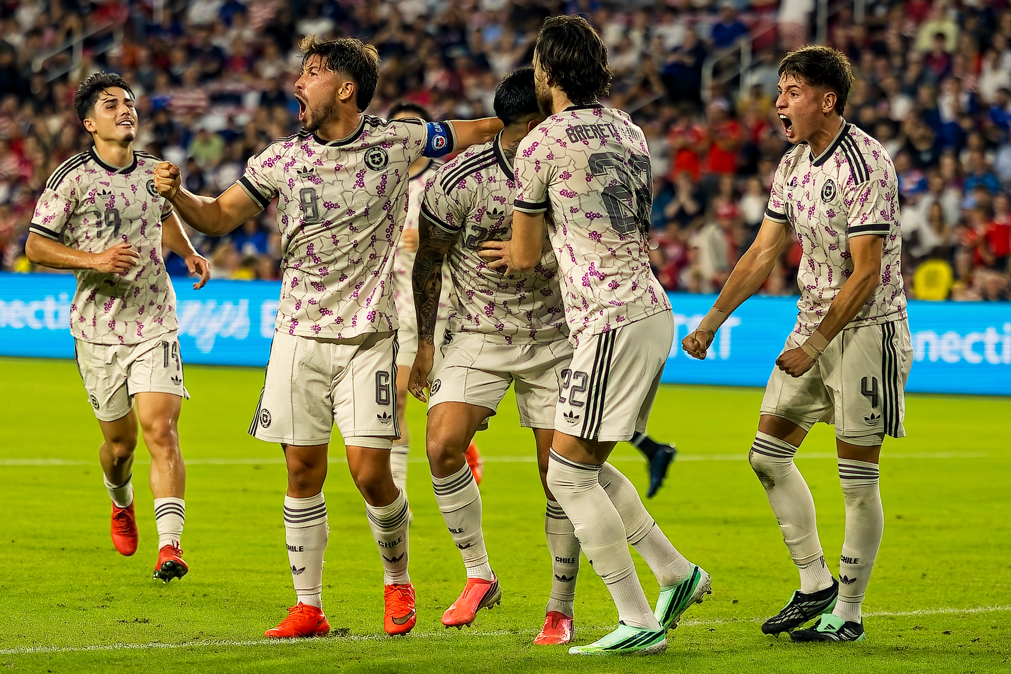 Five Chile players in white kits roar and embrace on the field while celebrating a goal, with the crowd blurred behind them.