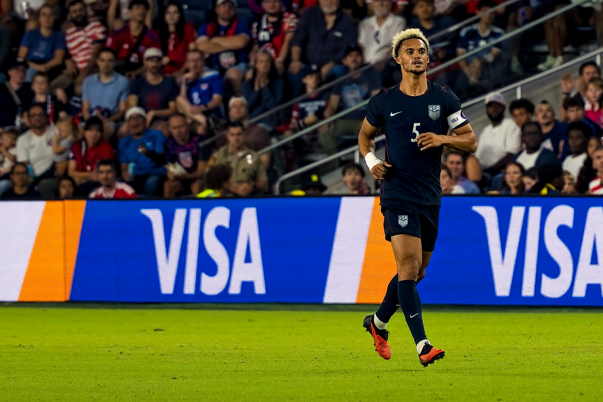 United States defender Antonee Robinson, wearing number 5 and the captain's armband, runs upfield in front of advertising boards and a full crowd.