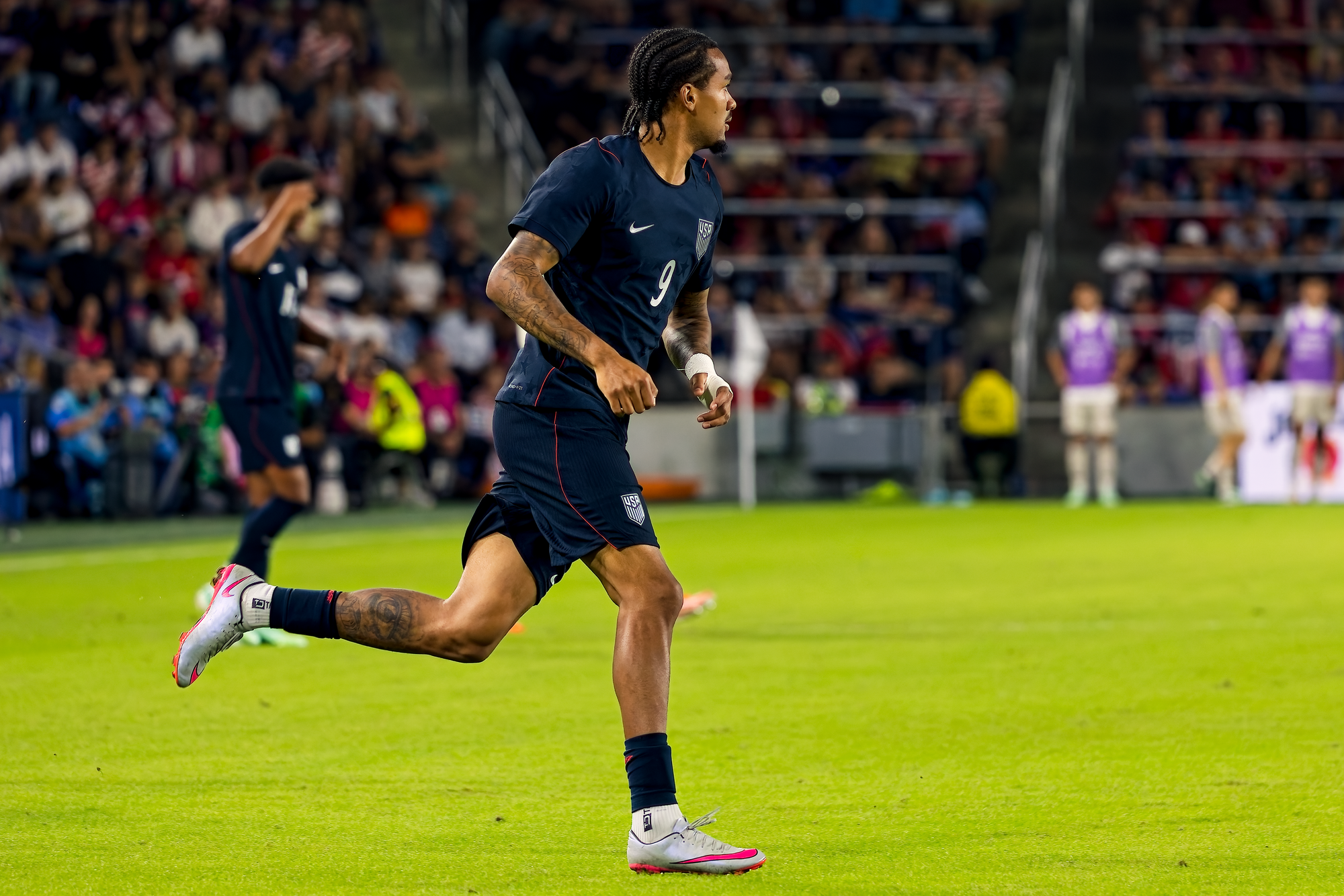 United States forward Damion Downs, wearing number 9, runs upfield with substitutes warming up on the sideline behind him.