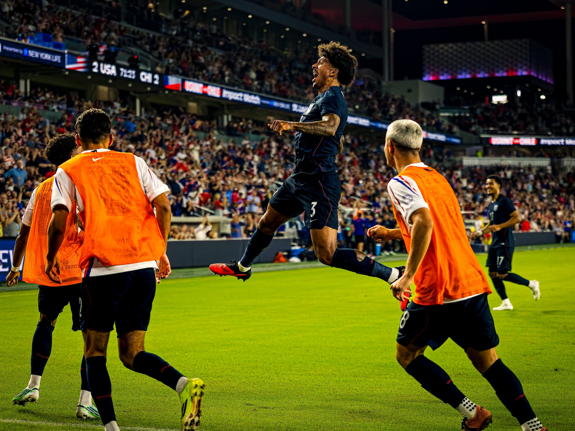 United States defender Chris Richards leaps into the air with his head back, shouting in celebration after scoring, as teammates in orange training bibs run toward him and the stadium scoreboard shows the match clock behind him.