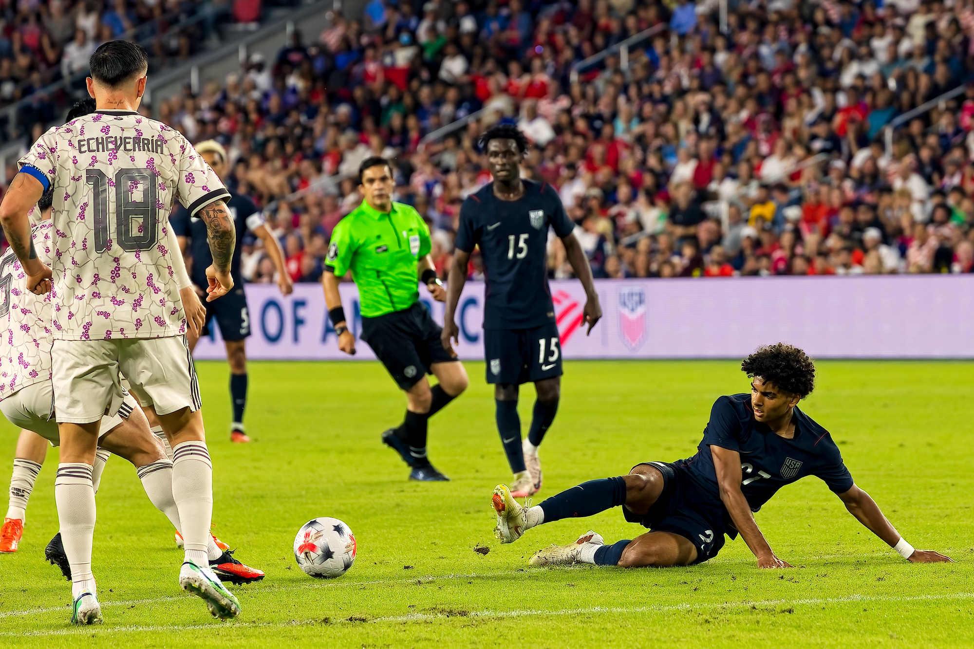 A United States player slides along the grass to challenge a Chile player who is playing the ball, with the referee and other players watching.