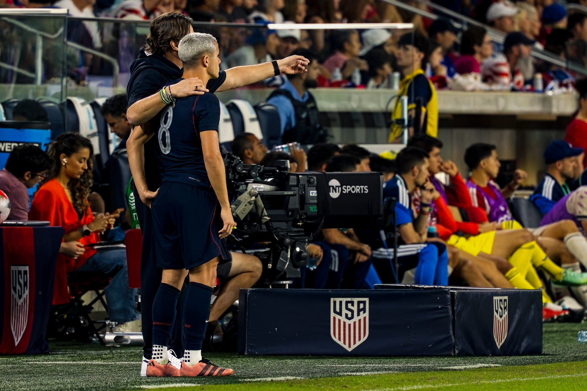 United States coach Mauricio Pochettino stands with his arm around substitute Cavan Sullivan at the touchline, pointing out onto the field as he gives instructions before the player enters the match.