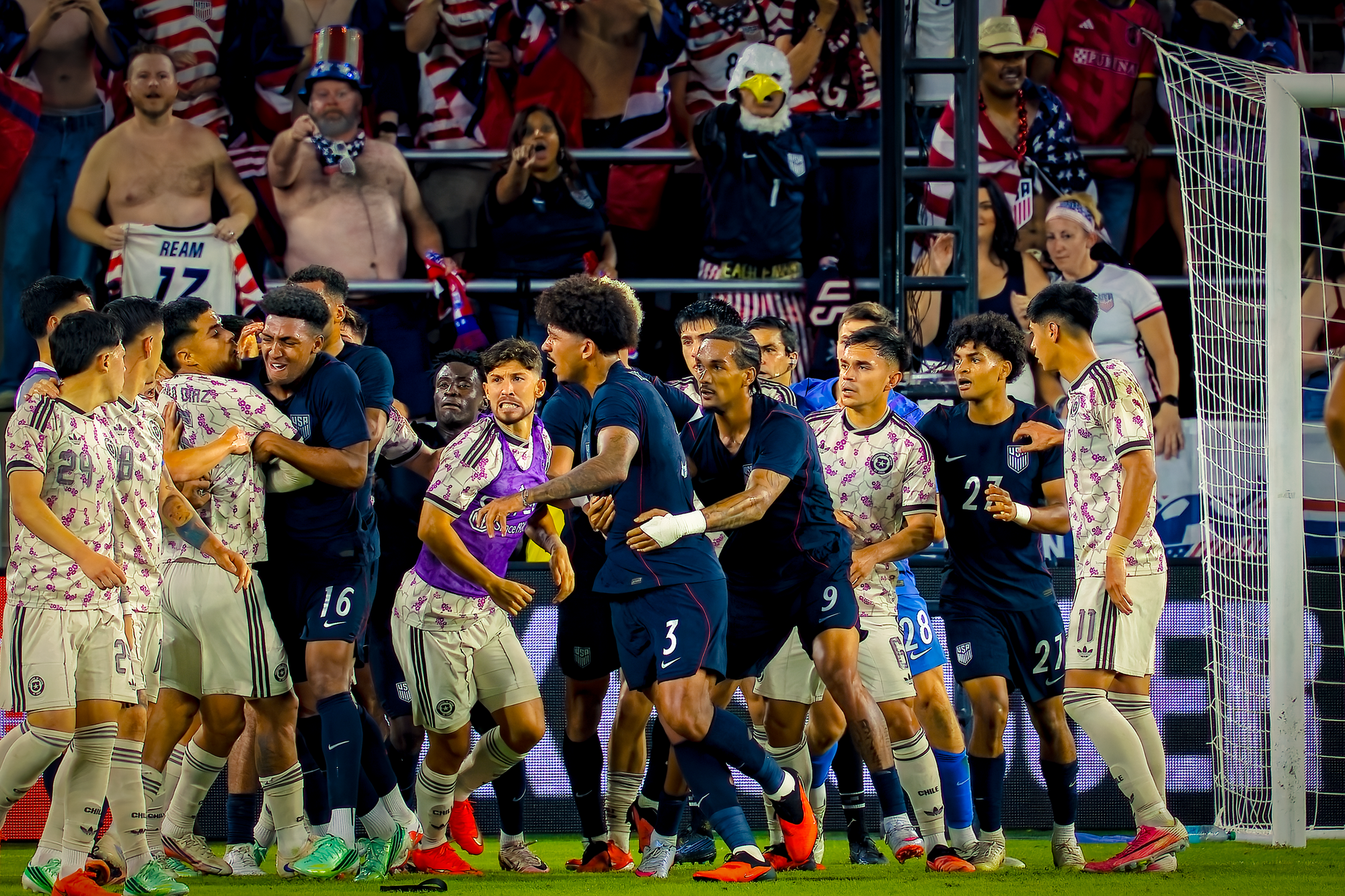 Players from both teams crowd together in a confrontation near the goal, grabbing at each other as fans look on from the stands behind.